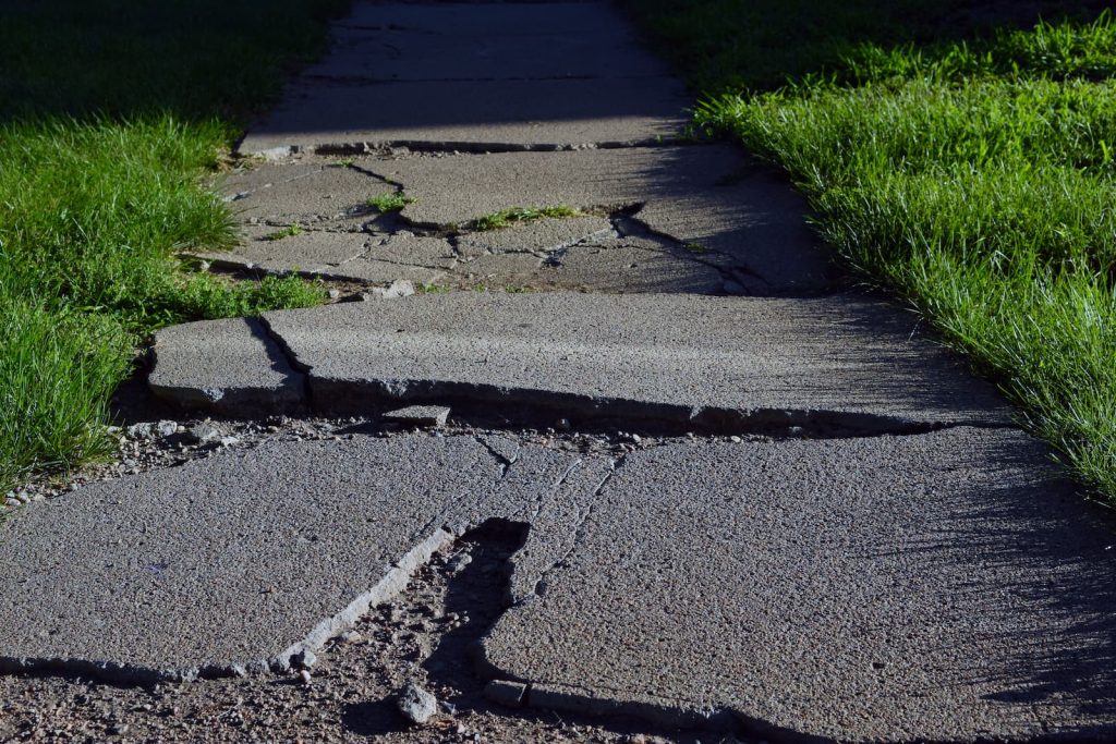 A close-up, low-angle view of a severely cracked and uneven concrete sidewalk with large gaps and lifted edges, flanked by green grass.